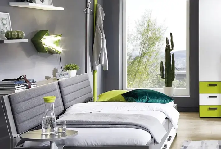 Graues Funktionsbett mit weisser Bettdecke und grünen Kissen, links Wandboard mit Lampe und Büchern, rechts grosses Fenster mit Kaktus und Kommode mit weissen und grünen Schubladen.